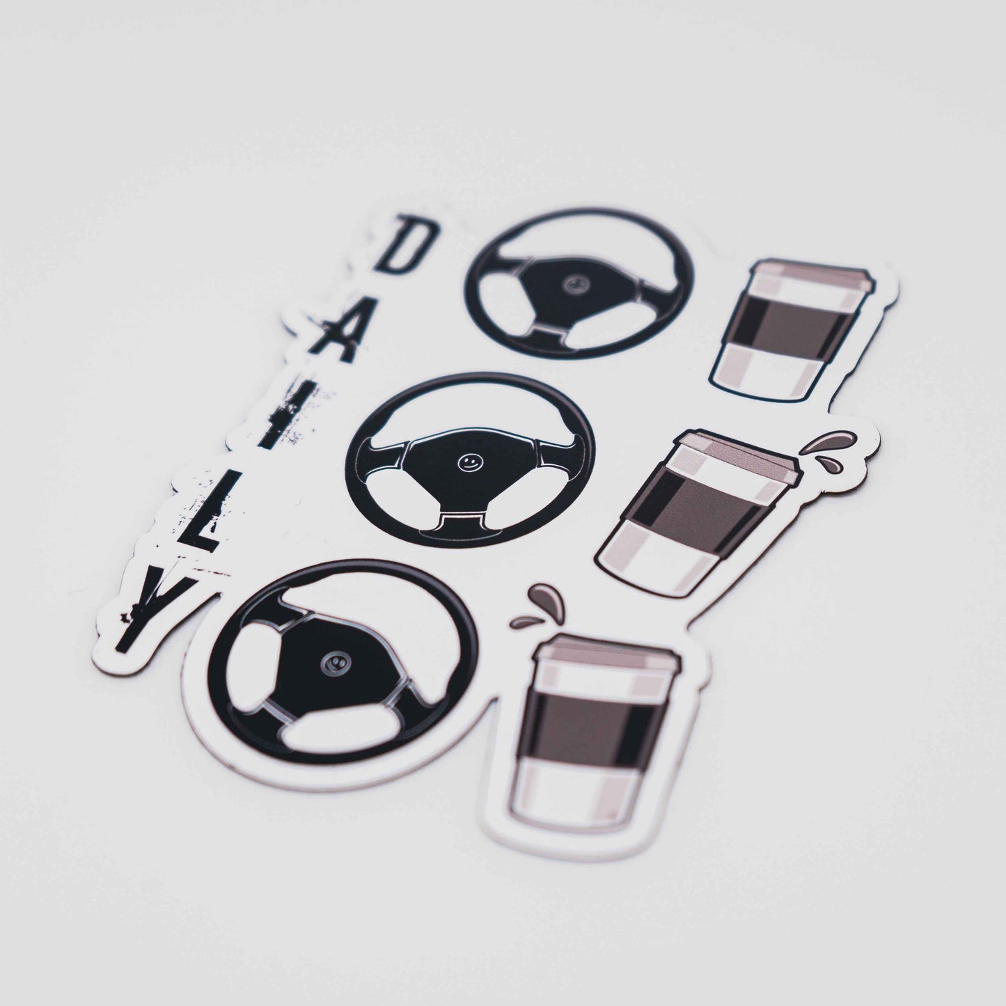 Closeup of the Daily-driver fridge magnet with a coffee cup tilting and splashing under g-force relative to the turning position of a car steering wheel over a white background. Smiley faces in the middle of the steering wheels that look in the direction the wheels are positioned indicate the joyful response of a driver.
